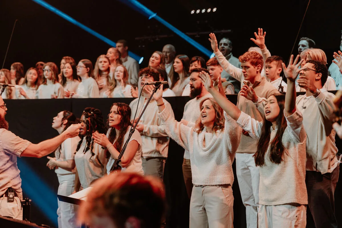 A large group of singers on stage with hands raised.